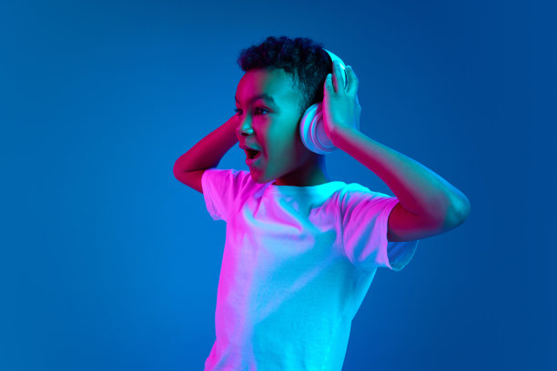 6-7岁的小男孩，穿着白色t恤，戴着耳机，在霓虹灯下听着蓝色背景的音乐。图片下载