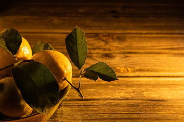 旁边的照片一碗充满新鲜和有机柠檬与他们的绿叶在桌子和黑色的背景。日落时强烈的侧光图片下载