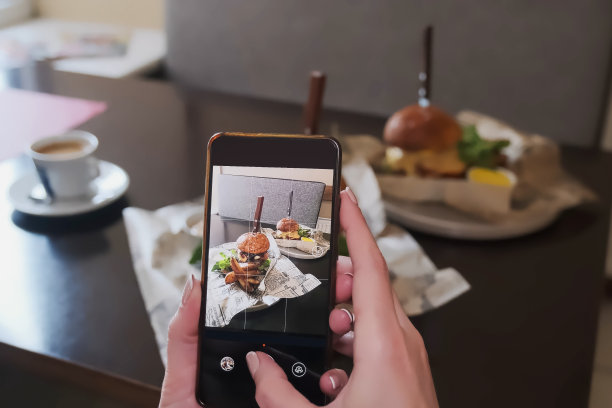 女人拍摄食物。女孩坐在餐厅或咖啡馆的桌子旁，用智能手机拍汉堡。第一个人的观点。手关闭图片下载