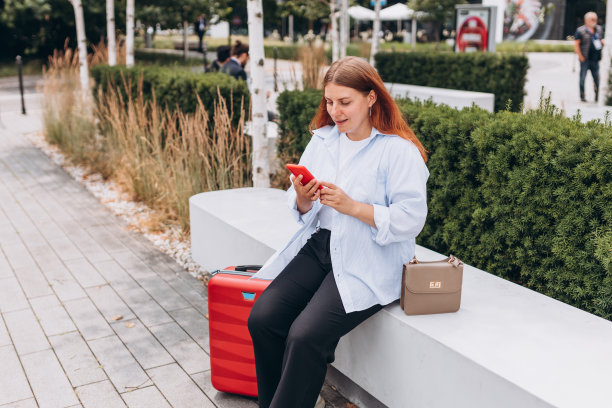 年轻的红发女子与红色行李箱使用智能手机在城市背景。快乐的女孩坐在户外的长凳上。城市生活理念，科技旗帜图片下载