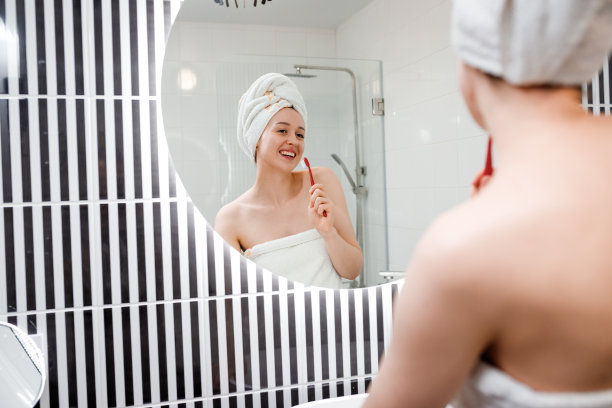 一个美丽的年轻女子穿着白毛巾在她的浴室镜子前刷牙的肖像。早上例行美容图片下载