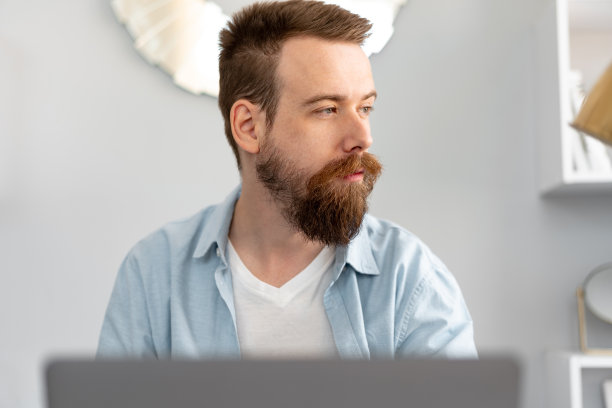 时髦的大胡子男人坐在家里的桌子上用笔记本电脑工作图片下载