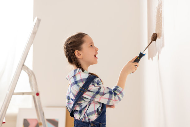 一个8岁的小女孩，穿着牛仔工装裤和格子衬衫，在她的育儿室里用油漆滚筒涂一堵棕色的墙。背景中有一架梯子图片下载