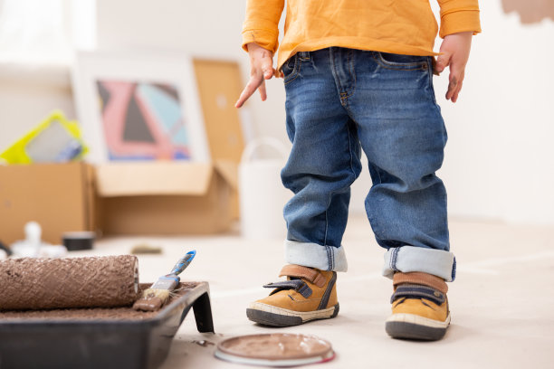 一个孩子的腿特写，穿着牛仔裤和橙色运动鞋。侧面是涂有棕色油漆的等离子碗。背景是需要修理的东西:盒子、画、罐子、画笔、滚筒等等图片下载