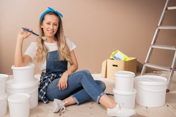 美丽的金发女人戴着蓝色头巾，穿着牛仔连体衣，坐在地板上，周围是用手刷的墙壁油漆容器。这位女士在搬家前正在自己装修公寓图片下载
