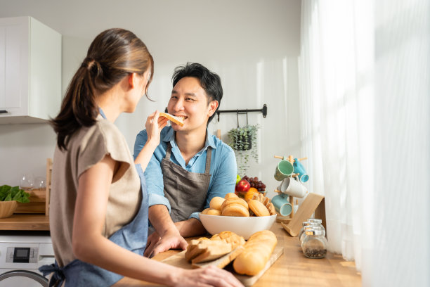 亚洲年轻女性拿着面包在厨房给男性。有魅力的男女夫妇穿着围裙，花时间在家里做早餐保健。饮食和健康食品的概念图片下载