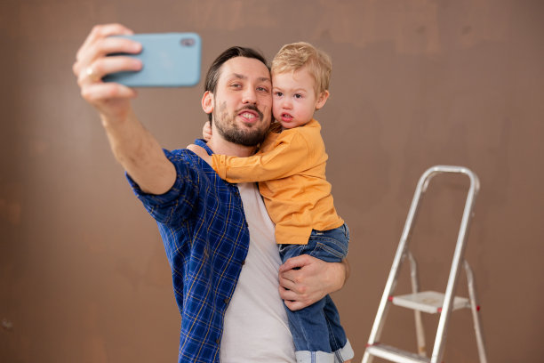 一位深褐色头发的父亲抱着他金发碧眼的儿子，在他们大楼装修开始的时候拍了一张纪念自拍照。那人的脸上涂满了颜料。图片下载