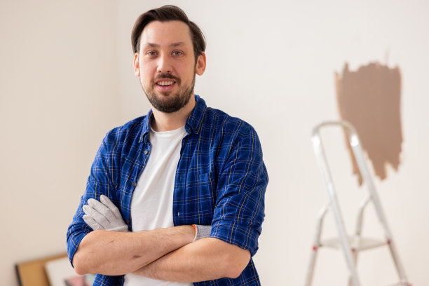 一个穿着白t恤和蓝色格子衬衫的英俊的褐发男子的特写。他微笑着，双臂交叉。他身后是一架梯子图片下载