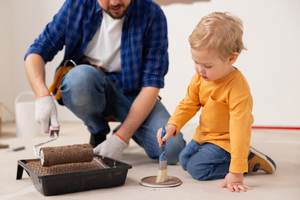 一个两岁的金发男孩穿着橙色毛衣和牛仔裤，用画笔在地板上作画。这个孩子非常专注于他正在做的事情。他的父亲在他身边图片下载
