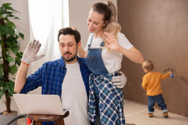 身穿牛仔布和格子衬衫的两名英俊的年轻人正在和朋友们视频聊天，向他们展示装修后的房子。背景是他们的儿子正在刷墙图片下载