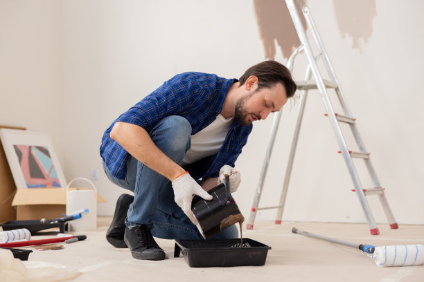 一个穿着牛仔裤、白t恤和格子衬衫的棕色头发的英俊男子正在把棕色油漆倒进一个等离子施工碗里。在背景中，他有一些表示翻新的东西图片下载