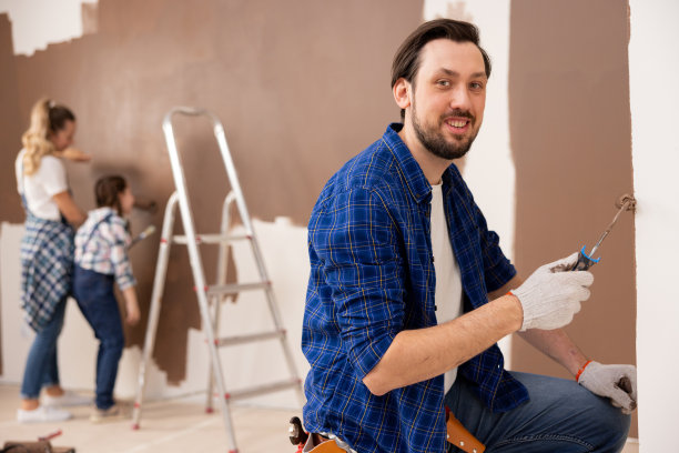 一个微笑着的黑发男人用一个小滚筒把墙涂成棕色。在他身后，从后面看，是他的妻子和女儿。他们都参与了维修工作图片下载