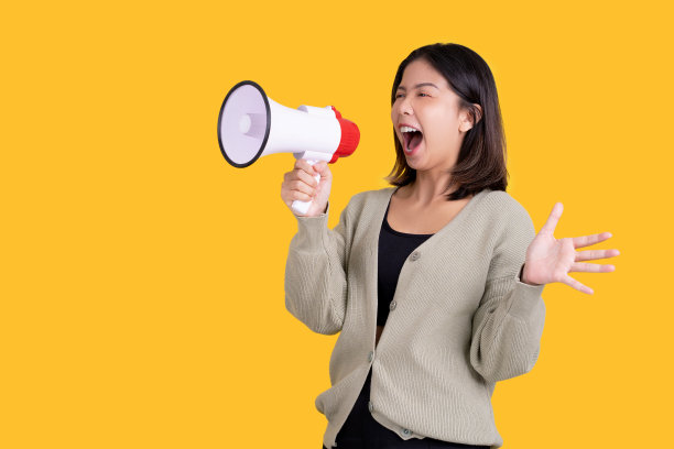 亚洲妇女在扩音器中尖叫，摊开她的手看向一个明亮的黄色背景。图片下载