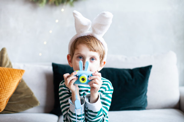 小男孩穿着条纹睡衣，戴着兔子耳朵的头巾，坐在沙发上，手里拿着给孩子们的兔子相机。图片下载