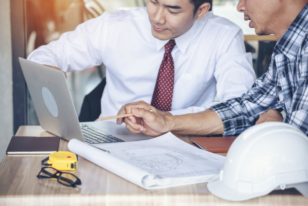 承包商施工工程师在施工办公室现场的建筑师桌子上开会交谈。商人、工程师、经理与工头讨论，团队建设者看计划蓝图笔记本电脑图片下载