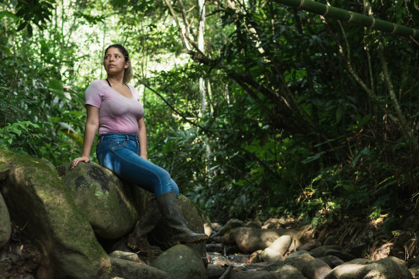 美丽的拉丁农妇，在哥伦比亚潮湿的丛林中，坐在一块石头上，带着好奇和严肃的表情向右边看。在南美洲探索美丽森林的女孩图片下载