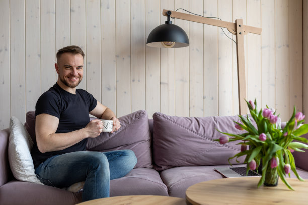 肖像微笑的男人拿着白色的咖啡杯，坐在柔软的紫罗兰色沙发上，穿着便装，靠近桌子的花瓶新鲜的郁金香。客厅设计优雅，装饰美观图片下载