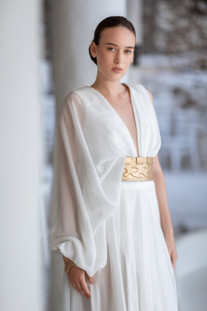 年轻漂亮的时尚模特身穿白色丝绸连衣裙，系着金色腰带，站在墙边，看着镜头图片下载