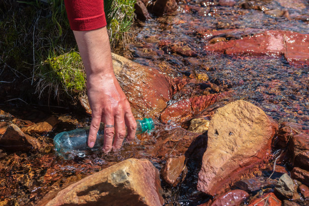 人类在明亮的阳光下从山溪中补充水。游客在阳光明媚的日子里往瓶子里装满矿泉水。手拿瓶子在阳光下的清水流。徒步时补充水分。图片下载