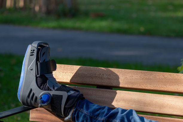 骨折后脚骨折的男孩和矫形鞋或助行器在公园长凳上休息，脚抬起来，用医疗设备和石膏重建和恢复他的痛苦事故图片下载