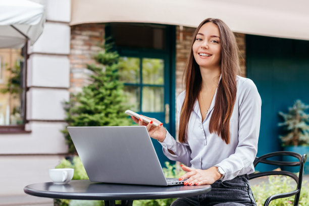 一个年轻的女学生坐在街边的咖啡馆里，用笔记本电脑和智能手机看着相机。远程工作，自由职业，在线教育图片下载