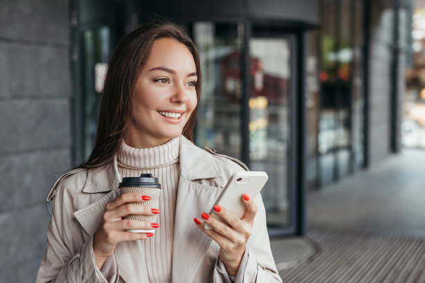 年轻的微笑的褐发女子拿着智能手机和一杯咖啡对着办公楼的背景，看向了另一边。本空间图片下载