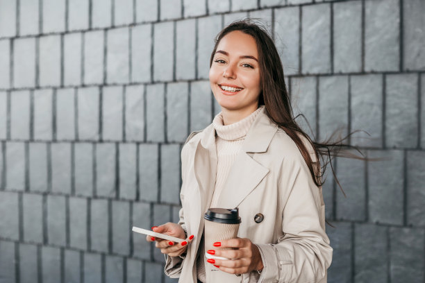 一个拿着智能手机的黑发女孩站在建筑正面的背景前微笑着。本空间图片下载