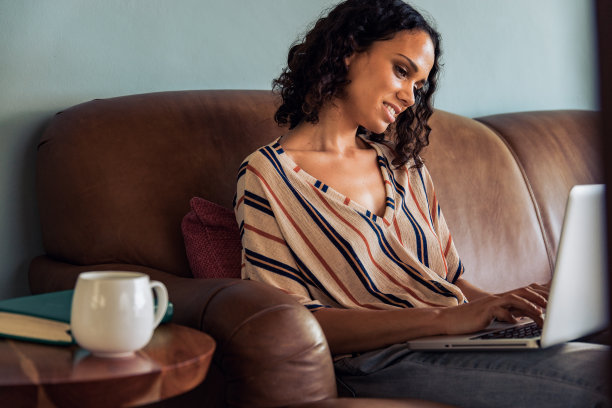 年轻女子坐在沙发上用笔记本电脑工作。非洲妇女在家里用笔记本电脑工作。图片下载