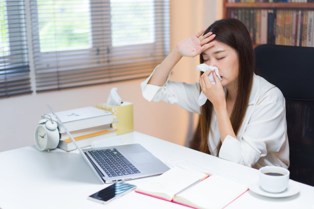 感觉又累又恶心。生病的年轻有吸引力的亚洲妇女与组织打喷嚏攻击，擤鼻子，而坐在她的工作地点在办公室。图片下载