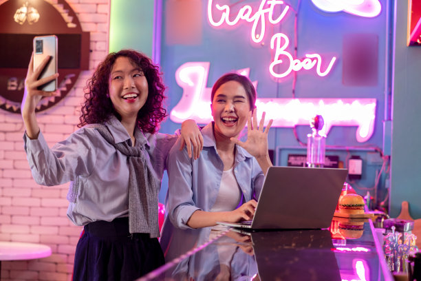 一位女商人在鸡尾酒吧用智能手机自拍，同时用笔记本电脑和同事讨论工作图片下载