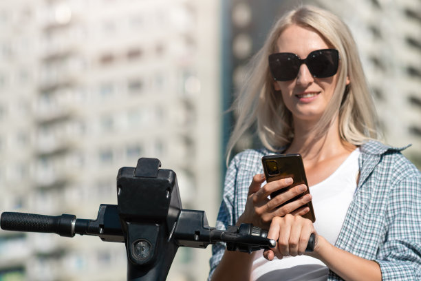 年轻微笑的金发女人戴着太阳镜，在一个阳光明媚的日子用手机打开一辆电动摩托车，复制空间图片下载