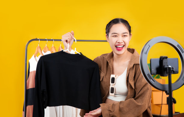 直播的概念，年轻的亚洲博主妇女展示衣服在智能手机摄像头前直播聊天和销售在她的网上商店在家里。图片下载