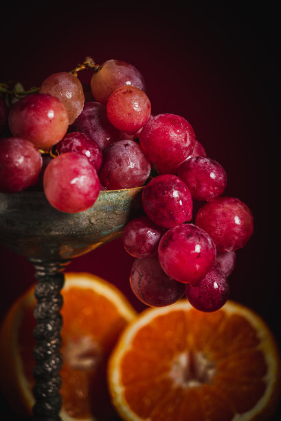 红色葡萄的细节在一个古董金属花瓶与两个橘子，在一个木制和红色的背景图片下载