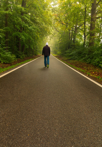 雾气蒙蒙的早晨，年轻人走在柏油路上的绿色森林里图片下载