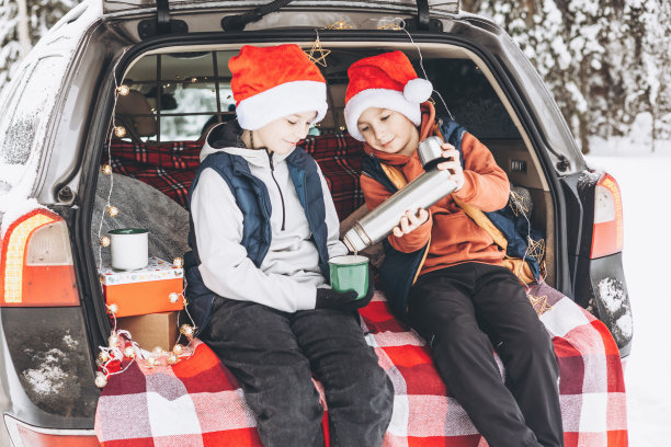两个朋友十几岁的男孩在红色的圣诞老人帽子与保温瓶坐在后备箱的汽车装饰圣诞节。自驾游冒险和当地旅游理念。冬季野餐在雪林新年假期图片下载