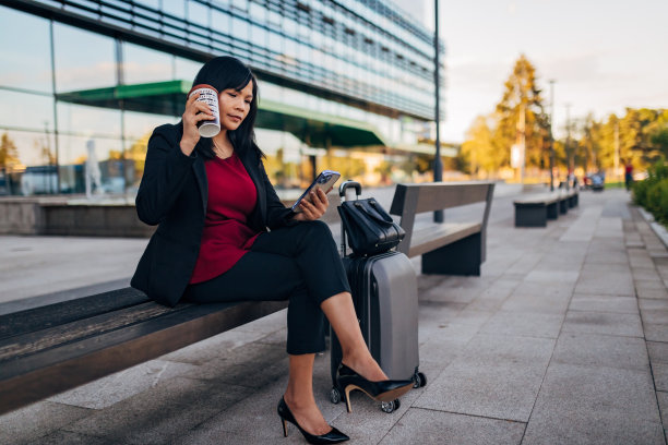 一位女商人坐在长凳上一边喝着外卖咖啡一边用手机图片下载