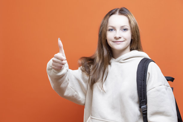 欧洲女十几岁的学生女孩与背包，手在口袋孤立的橙色背景与快乐的笑脸站大拇指向上的手势。水平射图片下载