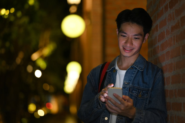 微笑的年轻亚洲男子站在夜晚的城市街道上用手机打短信。图片下载