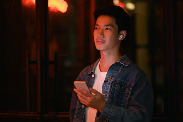 时髦的男人戴着无线耳机，用手机听音乐，走在夜晚的城市大道上。图片下载