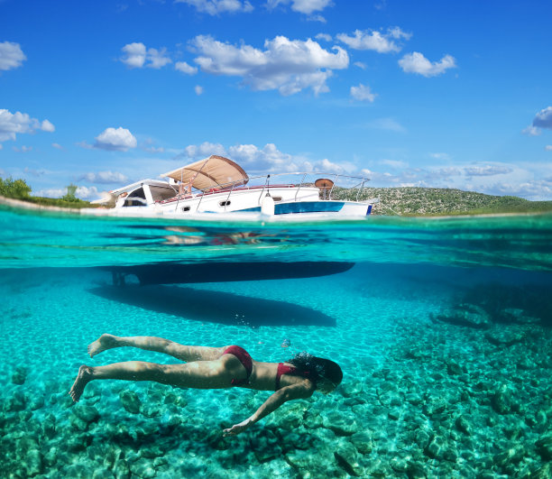 女潜水员正在美丽的海滩上浮潜。照片的下半部分是海底，上半部分是海岸，有一艘游艇和美丽的天空。图片下载