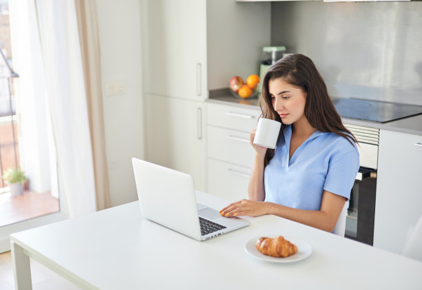 年轻女子一边吃早餐一边在家里使用笔记本电脑图片下载