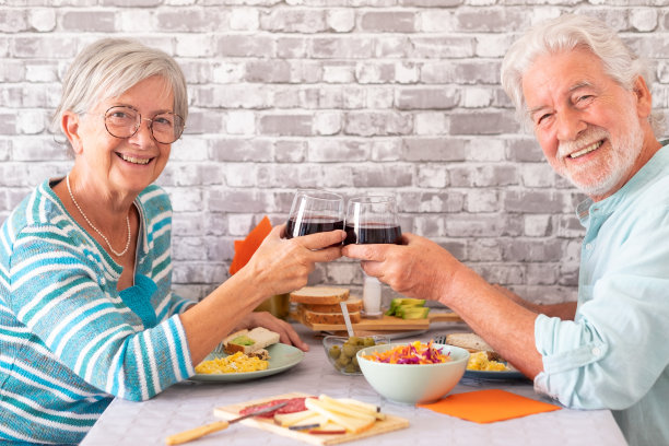 笑容满面的白人老夫妇在家里面对面坐在餐桌前吃早午餐，用红色的葡萄酒杯敬酒图片下载