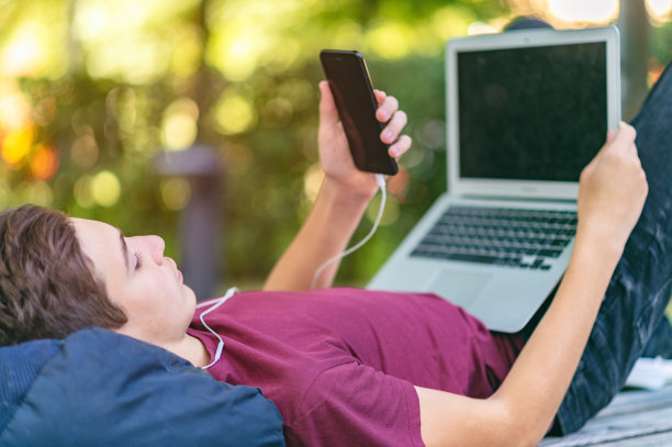 一个带着智能手机和笔记本电脑的少年躺在公园的长凳上。十几岁的男孩在户外打电话。白种少年穿着便装，在公园里拿着手机和电脑。图片下载