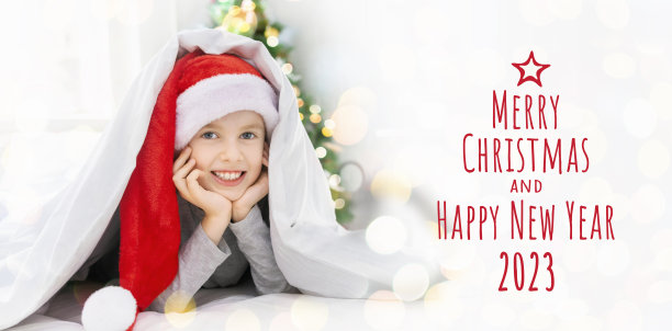 早上，戴着红色圣诞帽、盖着毯子的男孩微笑着躺在圣诞树前的床上。孩子看着镜头。背景文字圣诞快乐和新年快乐2023。图片下载