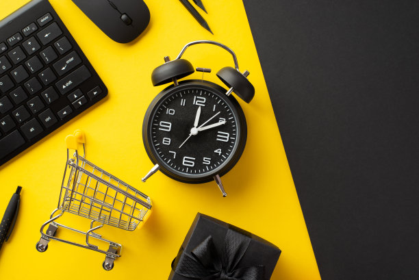 “黑色星期五”的概念。顶视图照片闹钟键盘电脑鼠标手推车铅笔和礼品盒与缎带蝴蝶结双色黄和黑色背景图片下载