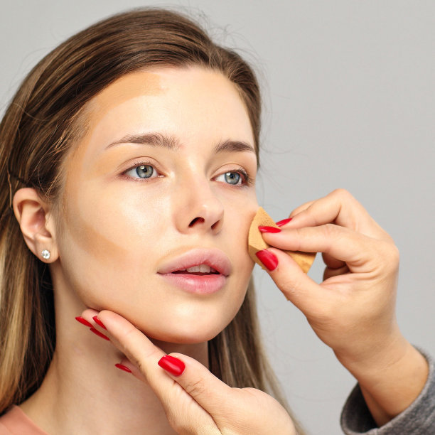 化妆师或造型师用湿海绵在女模特的脸上涂粉底。在脸上涂抹遮瑕膏。图片下载