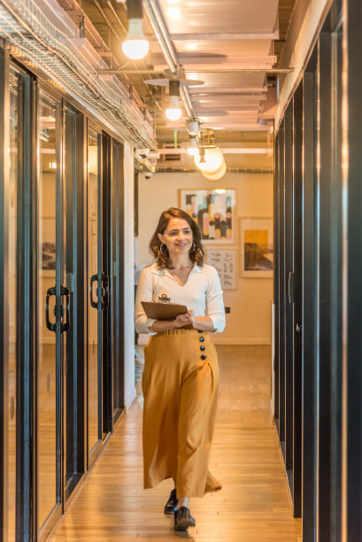 微笑着的年轻成年女性，穿着长裙，手里拿着积木，走在办公室走廊上图片下载