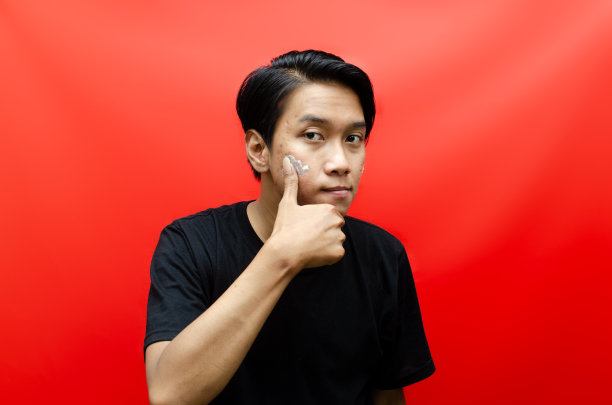 迷人的亚洲男子在红色背景的痤疮脸上涂抹面霜。图片下载