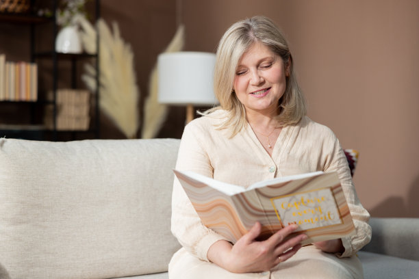一位美丽的老妇人面带微笑，拿着相册坐在客厅舒适的沙发上，开心地看着假期的家庭照片图片下载