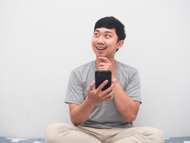 年轻积极的男人坐在床上拿着智能手机想着购物图片下载